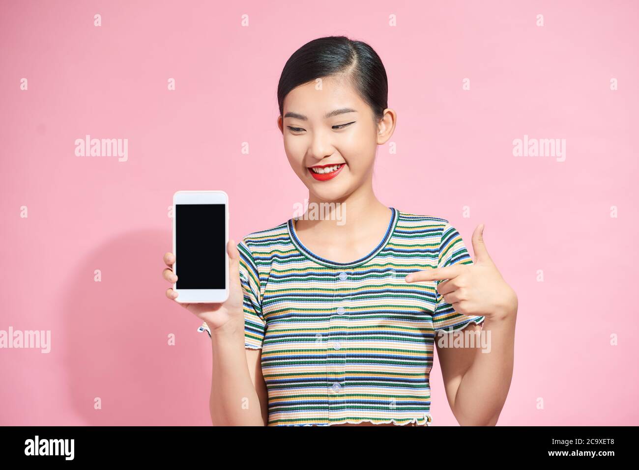 Ritratto di una ragazza asiatica carina che punta un dito su uno schermo mobile vuoto, isolato su sfondo rosa. Foto Stock