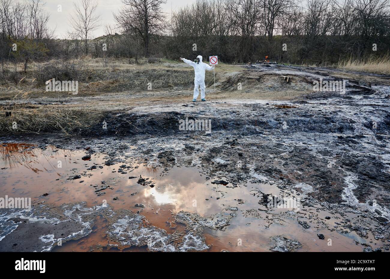Ricercatore in tuta protettiva bianca e maschera a gas che installa un cartello di avvertenza, nessun cartello d'ingresso, sul territorio contaminato. Acqua fangosa sporca arrugginita per analisi. Concetto di disastro ecologico Foto Stock