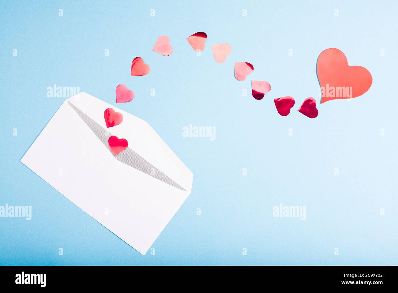 I cuori di confetti rossi escono da una busta bianca su uno sfondo azzurro. San Valentino. Concetto di amore dono, messaggio per l'amante Foto Stock