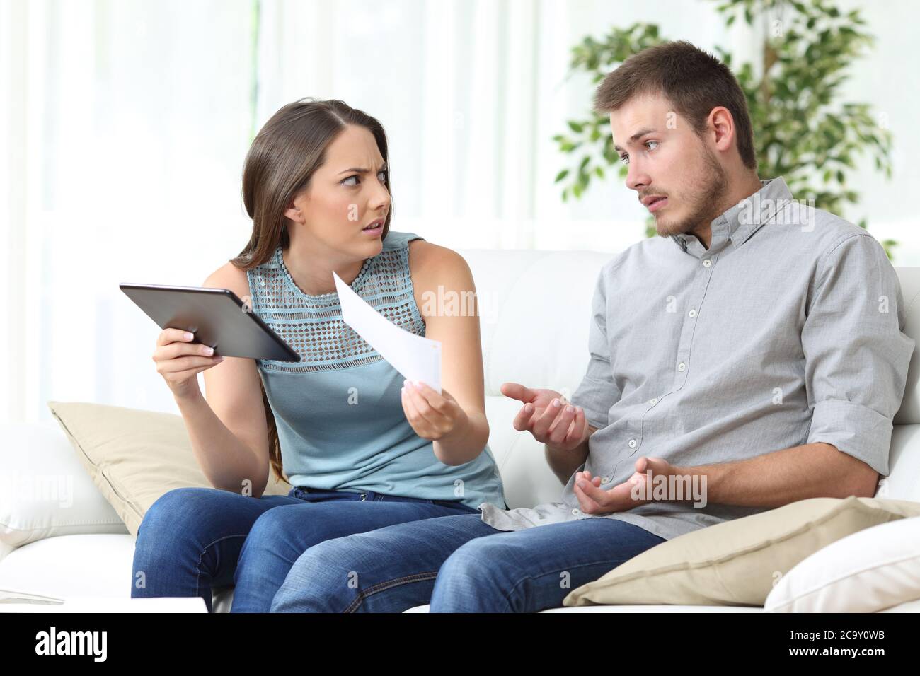 Moglie arrabbiata con un tablet che mostra ricevuta al marito seduto nel divano nel soggiorno di casa Foto Stock