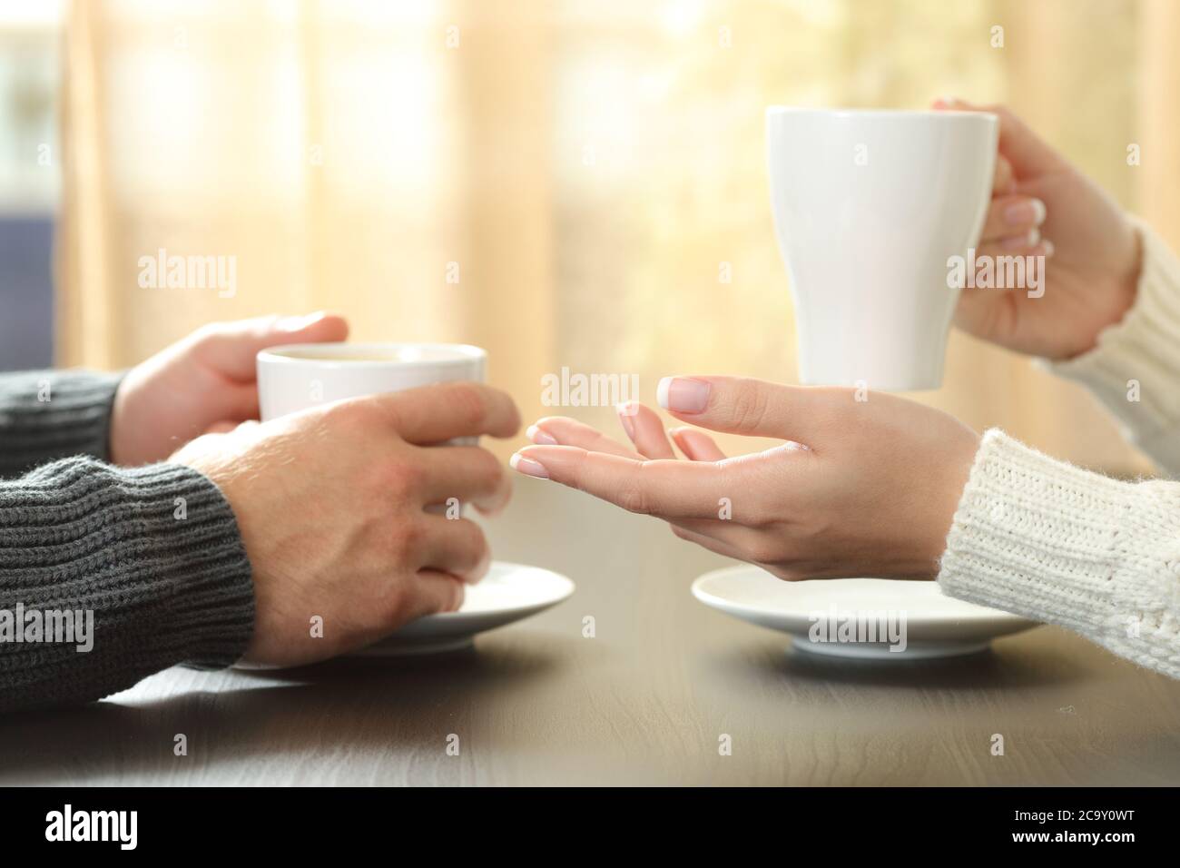 Primo piano di un paio di mani che parlano tenendo le tazze di caffè seduti su un tavolo accanto alla finestra a casa Foto Stock