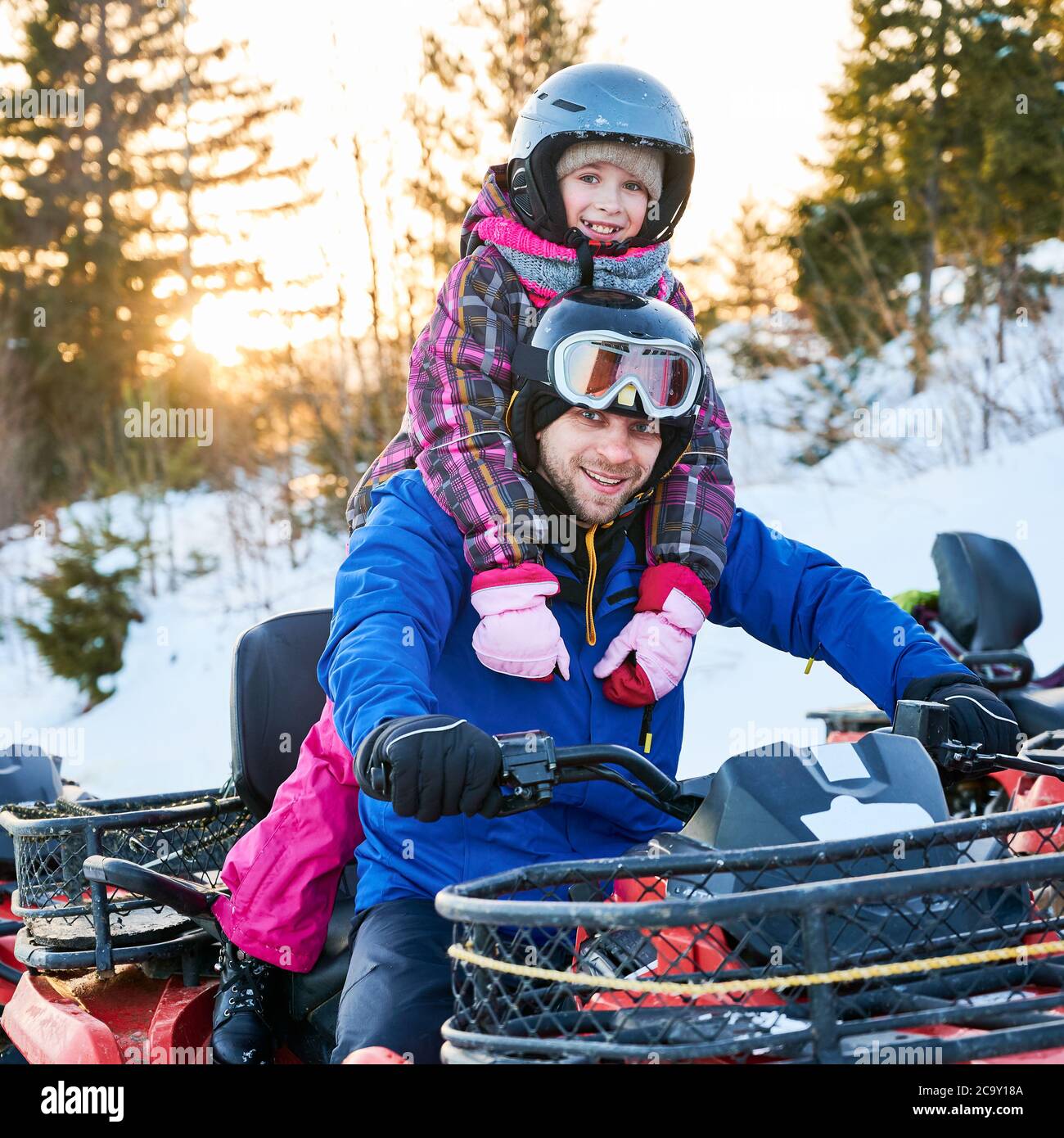 Ritratto di famiglia sorridente, padre e figlia, indossando vivaci costumi da sci, caschi e occhiali di protezione, seduto su una moto di squadra, sorridendo a macchina fotografica in montagne innevate al tramonto. Concetto di svago attivo. Foto Stock