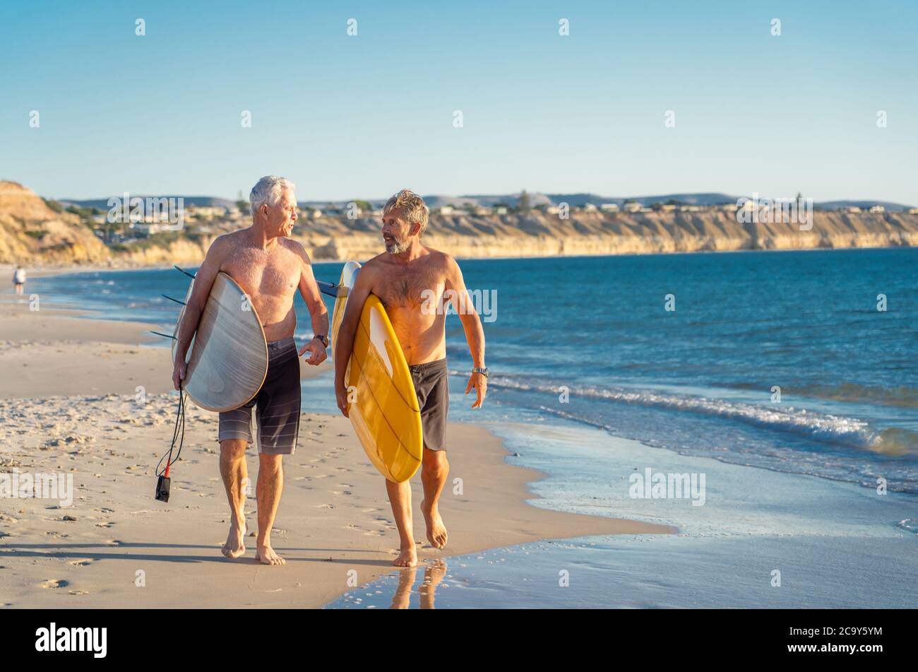 Due uomini maturi che camminano con tavole da surf sulla bellissima spiaggia godendo di paradiso e di vita di riposo. Amici adulti in forma attraente che si divertono Foto Stock