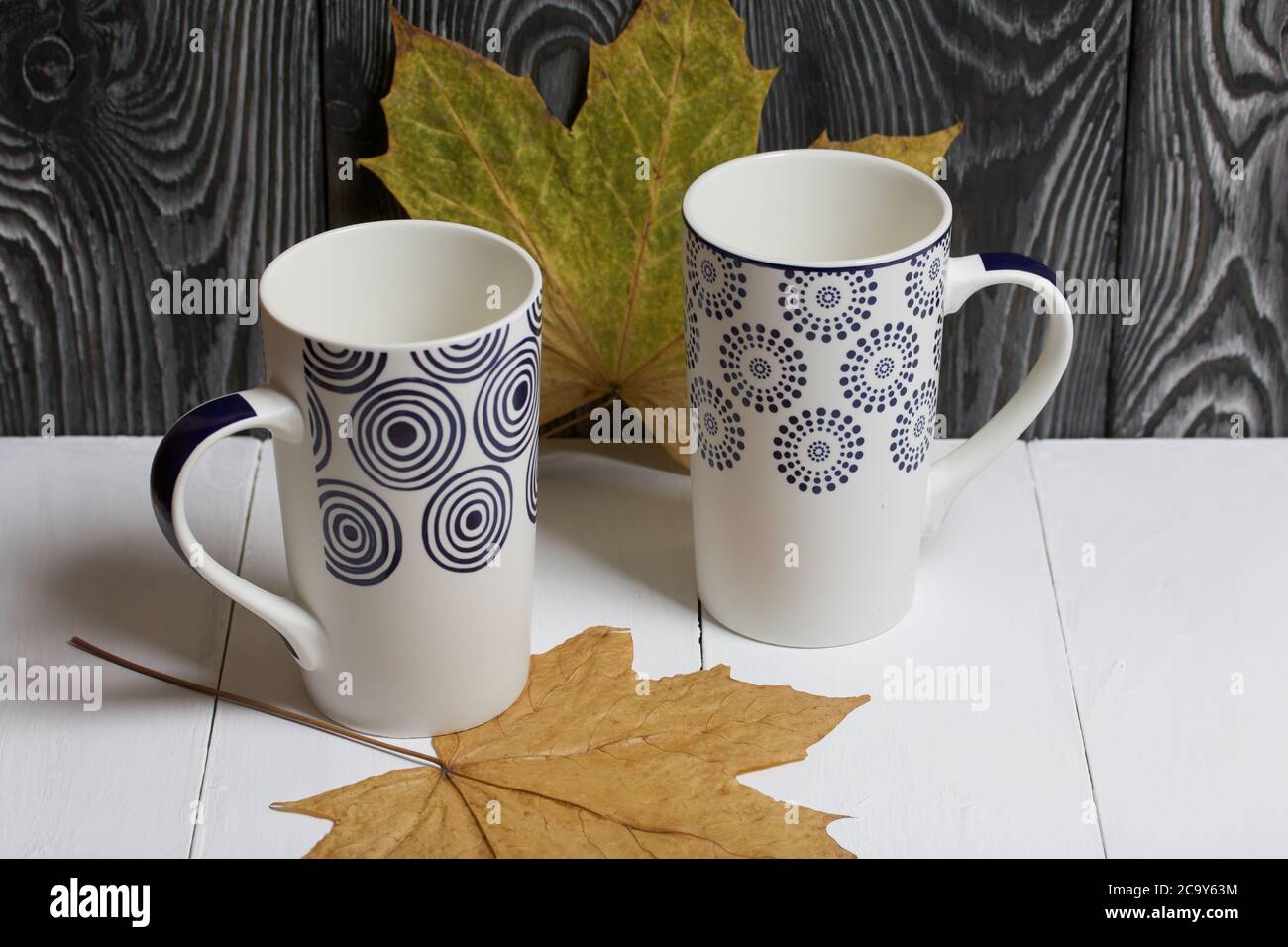 Tazza in ceramica su una superficie bianca. Tra di loro sono essiccate foglie di acero autunno. Su uno sfondo di tavole di pino dipinte di nero. Foto Stock