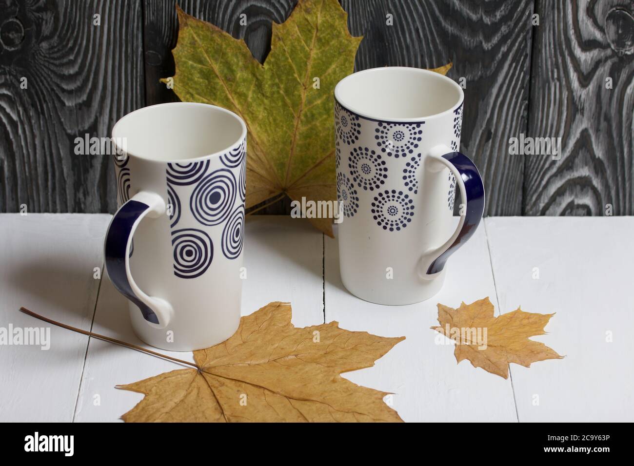Tazza in ceramica su una superficie bianca. Tra di loro sono essiccate foglie di acero autunno. Su uno sfondo di tavole di pino dipinte di nero. Foto Stock