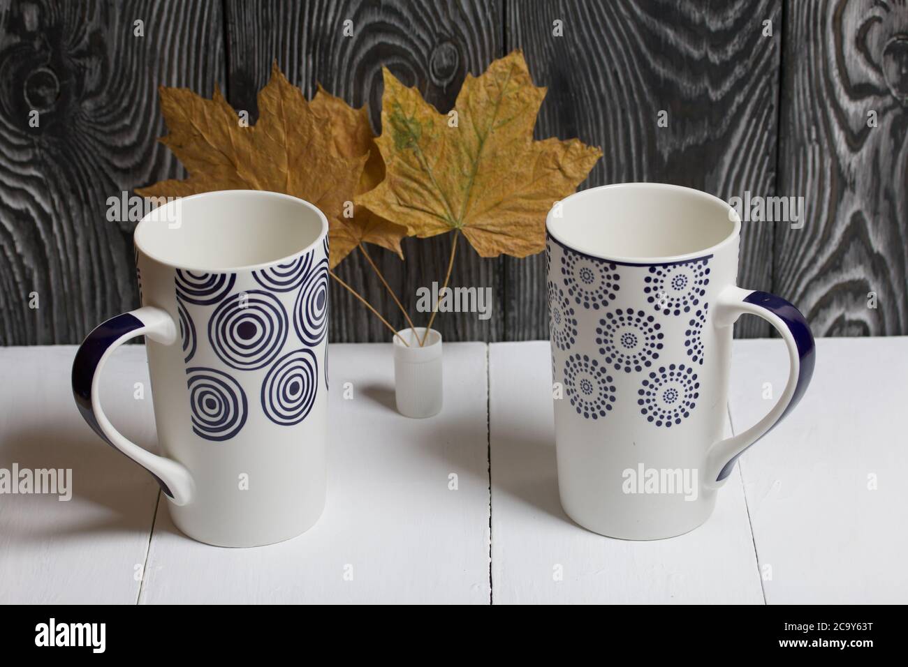 Tazza in ceramica su una superficie bianca. Tra di loro sono essiccate foglie di acero autunno. Su uno sfondo di tavole di pino dipinte di nero. Foto Stock