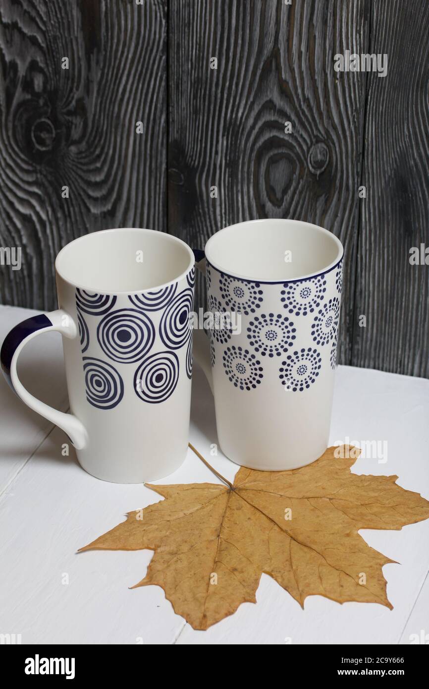 Tazza in ceramica su una superficie bianca. Tra di loro sono essiccate foglie di acero autunno. Su uno sfondo di tavole di pino dipinte di nero. Foto Stock
