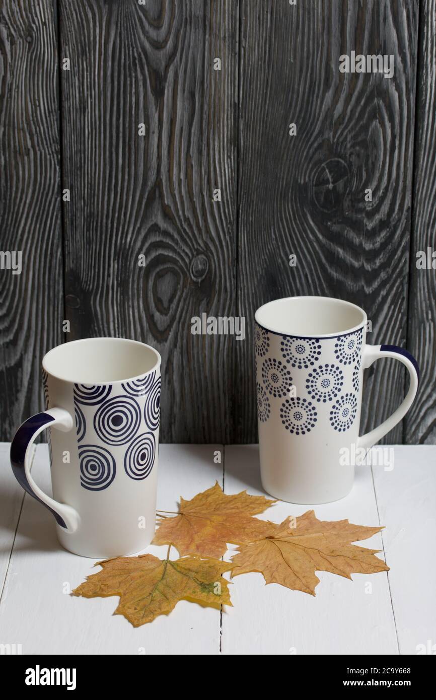 Tazza in ceramica su una superficie bianca. Tra di loro sono essiccate foglie di acero autunno. Su uno sfondo di tavole di pino dipinte di nero. Foto Stock