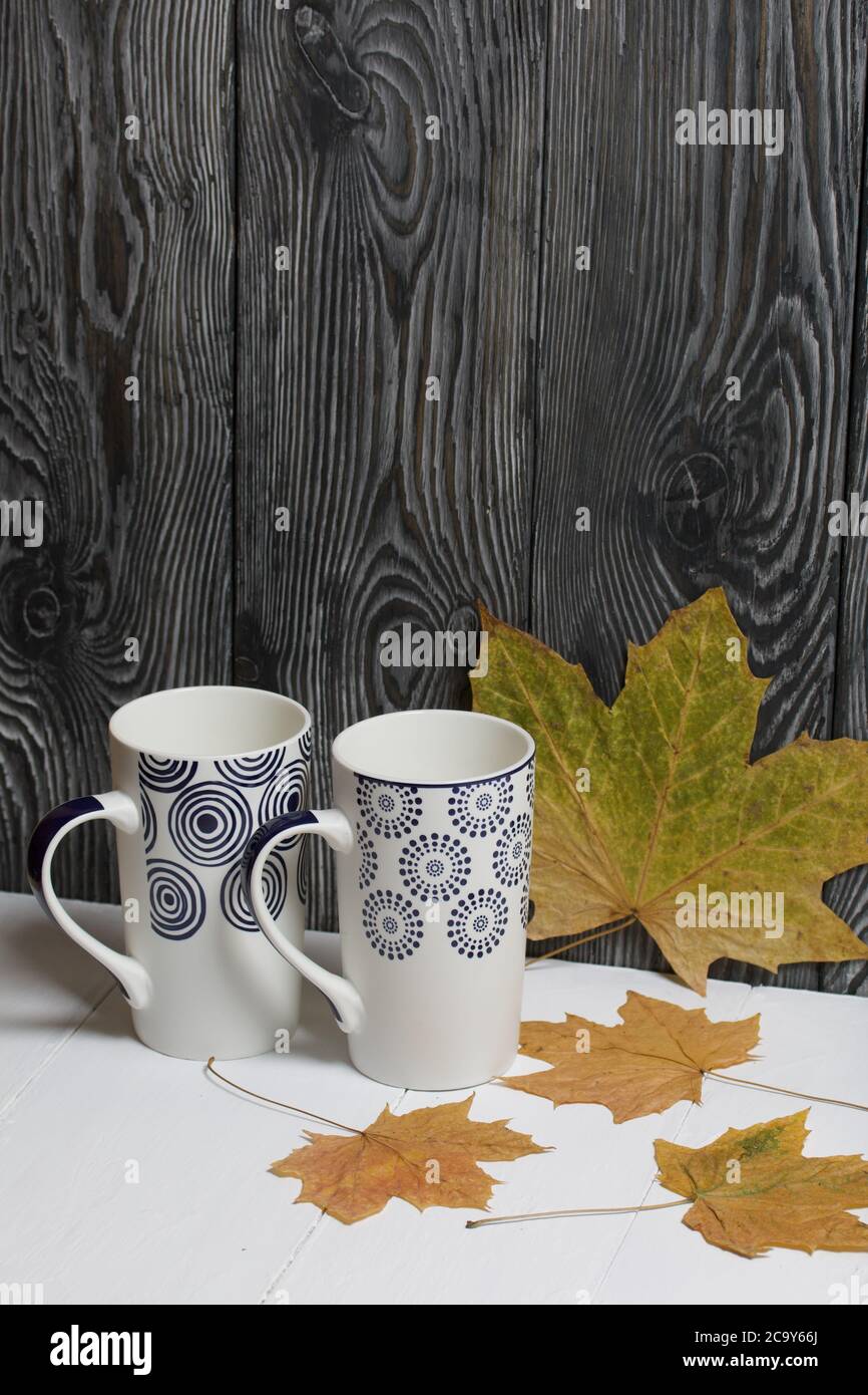 Tazza in ceramica su una superficie bianca. Tra di loro sono essiccate foglie di acero autunno. Su uno sfondo di tavole di pino dipinte di nero. Foto Stock