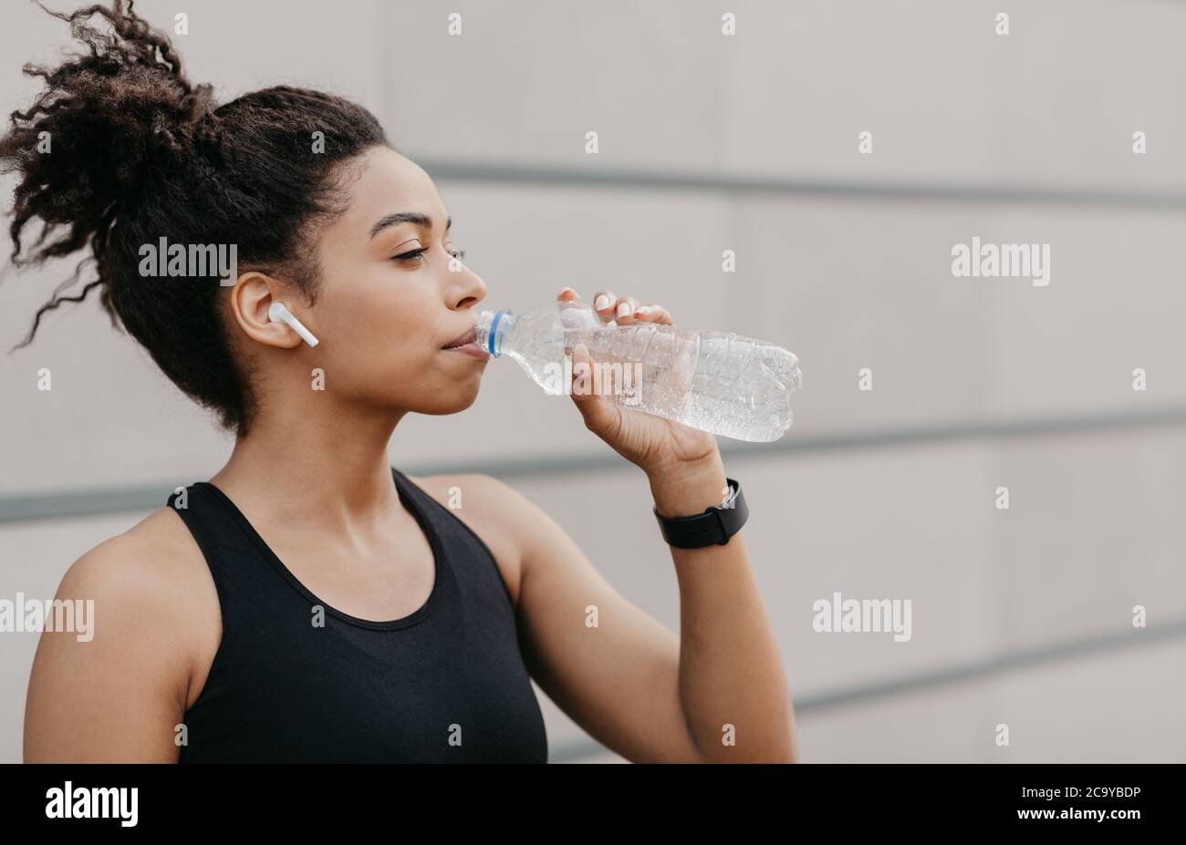 Croce, allenamento cardio e stile di vita sano. Muscolosa afroamericana ragazza con moderne cuffie e fitness tracker bere acqua Foto Stock