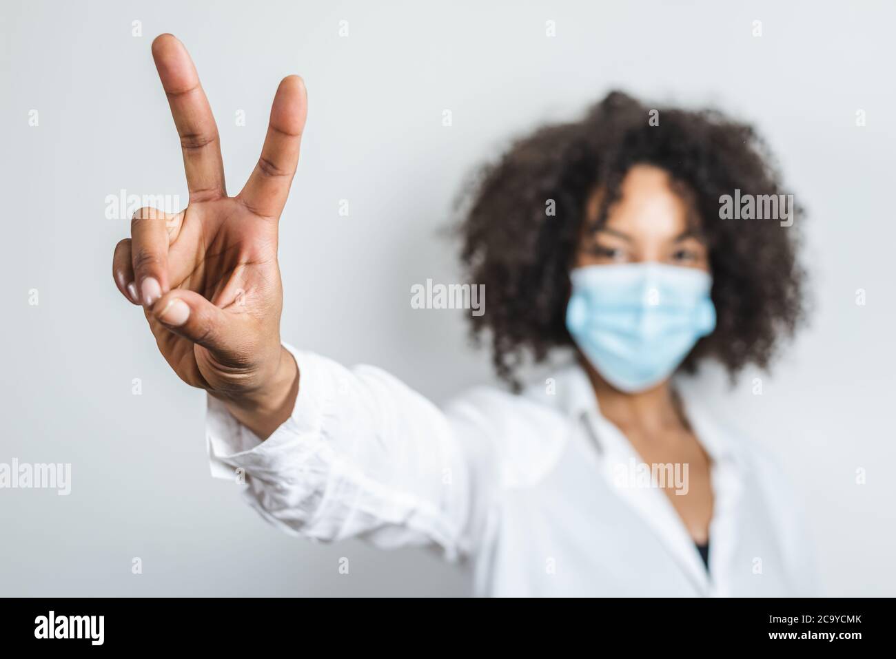 mano destra di una giovane donna afro usando una maschera chirurgica e facendo simbolo di vittoria con le dita Foto Stock