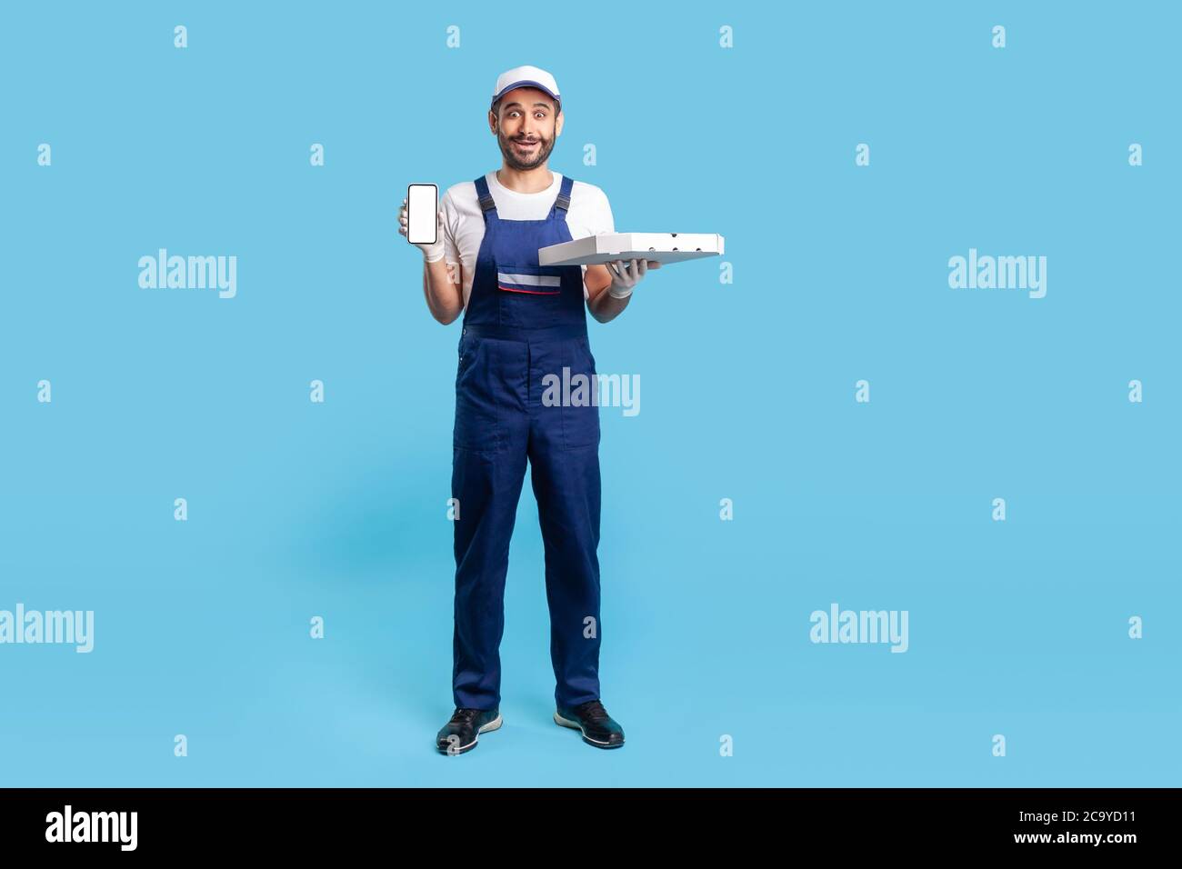 A lunghezza intera ha scioccato il corriere sorpreso in workwear e guanti di sicurezza che trasportano la scatola della pizza, mostrando il telefono mobile con il mock-up display per l'immagine di pubblicità. Foto Stock