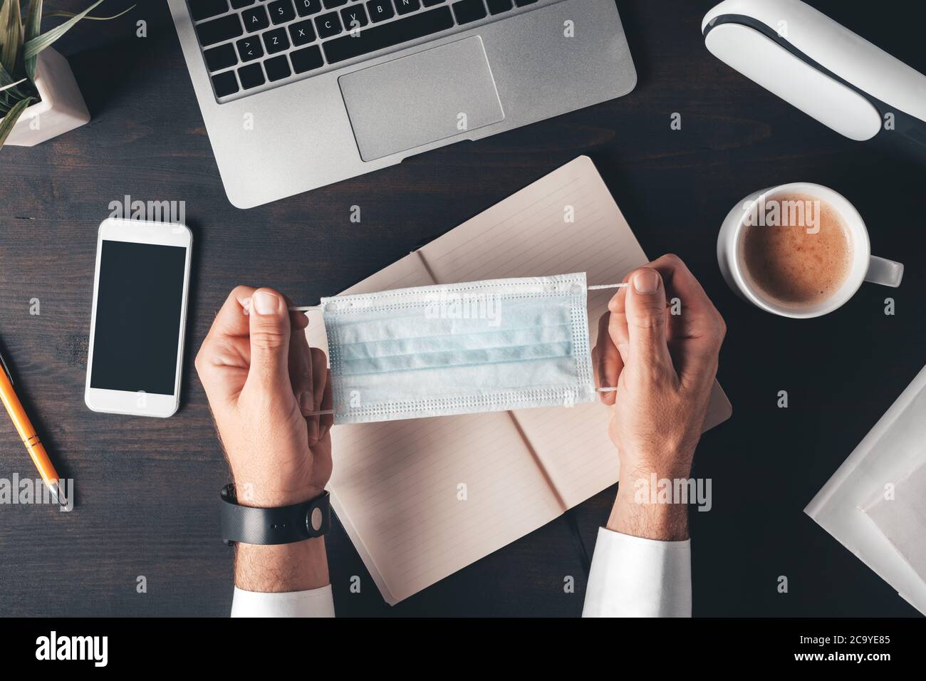 Uomo d'affari che tiene la maschera respiratoria protettiva in ufficio, vista dall'alto delle mani maschili sopra la scrivania Foto Stock