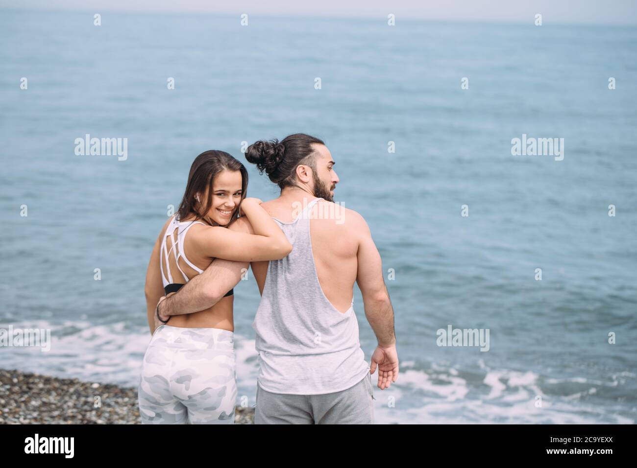 Giovane fitness coppia attrattiva in outfit da corsa, rilassante dopo un intenso allenamento, abbracciando sulla spiaggia, guardando all'orizzonte Foto Stock