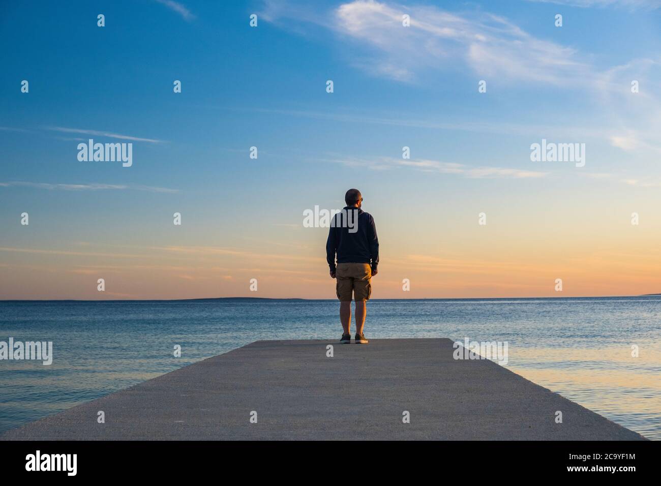 Uomo in felpa con cappuccio e pantaloni da carico corti in piedi sul molo e guardando all'orizzonte del mare, isola d Pag, mare Adriatico, Croazia Foto Stock