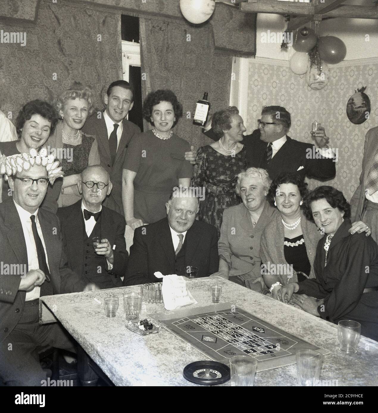 1960, storico, festa di compleanno per adulti in una casa, uomini e donne ben vestiti che festeggiano con alcune bevande, gin e un gioco di roulette da tavolo, con una piccola ruota e tavola seduti su un tavolo coperto di stoffa sala da pranzo, Inghilterra, Regno Unito. Foto Stock