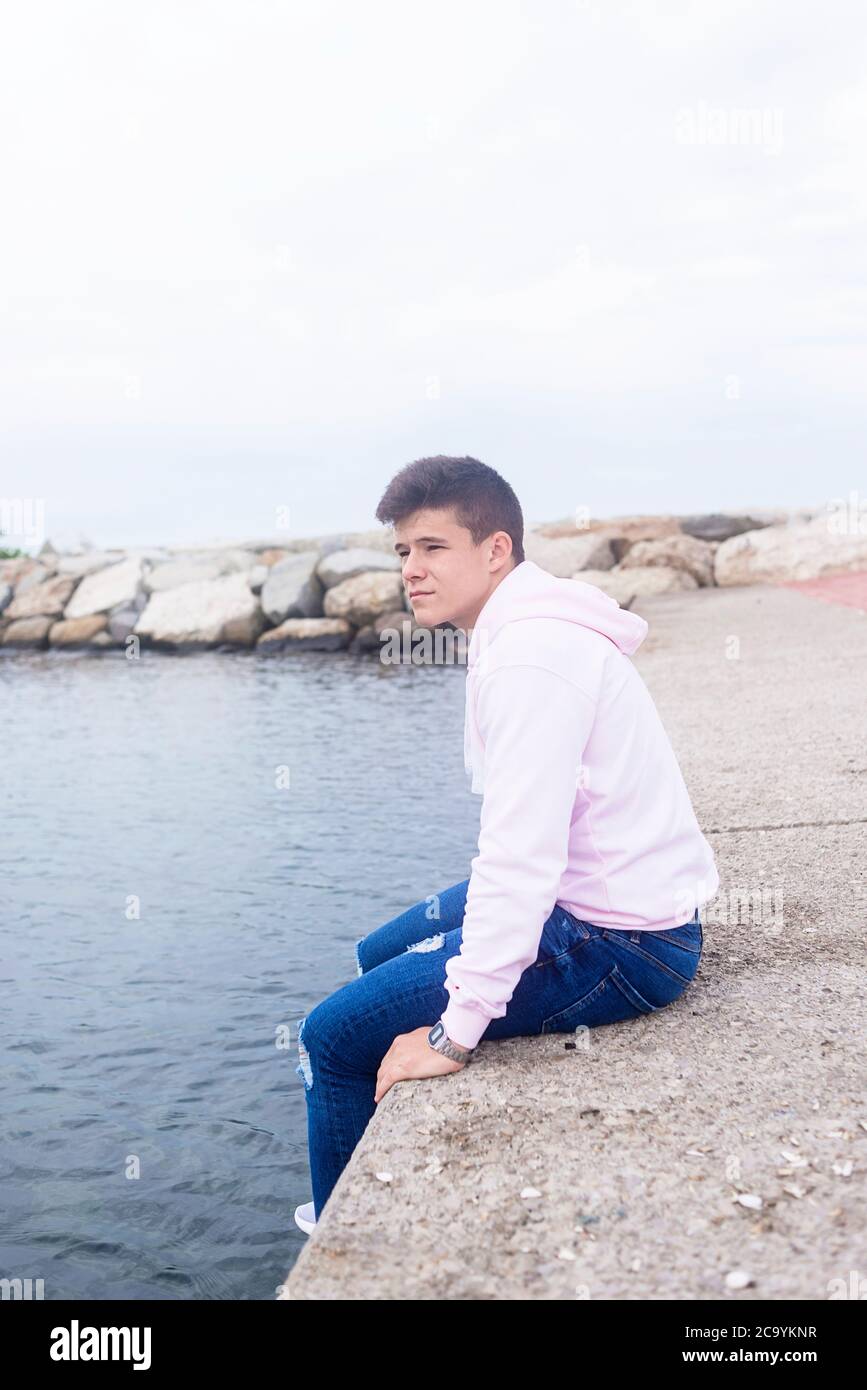 Giovane adolescente pensivo seduto su frangiflutti mentre si guarda la macchina fotografica Foto Stock