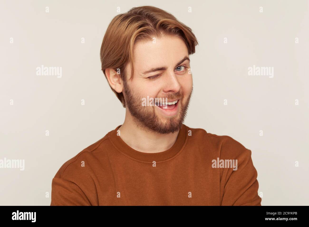 Ritratto di un giovane ottimista felice con capelli e barba puliti che indossa una felpa che si inaridisce alla macchina fotografica, guardando giocosamente e occhi lampeggianti, rallegrandosi. Foto Stock