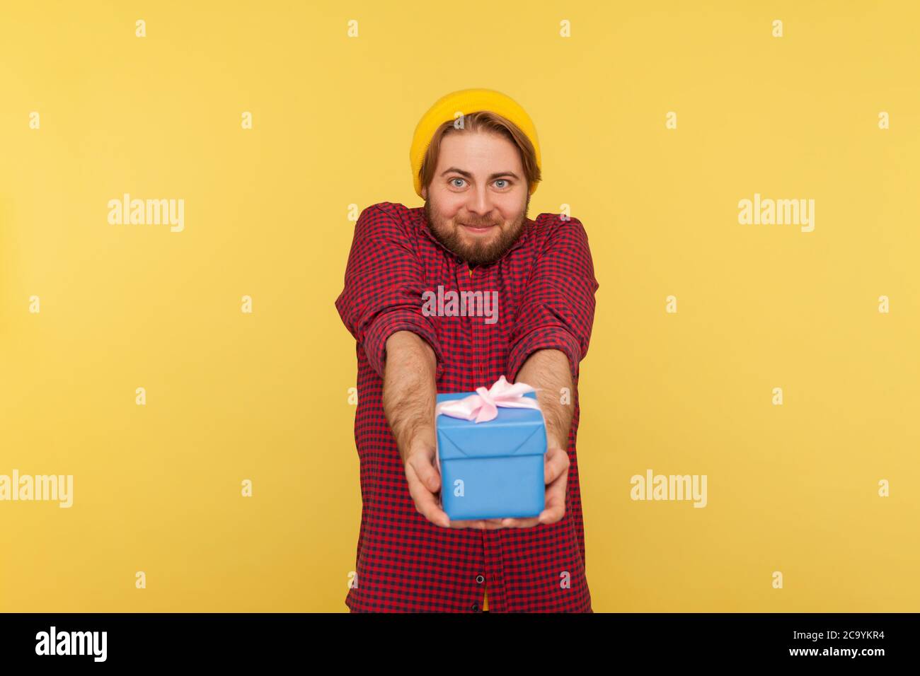 Tipo positivo generoso hipster ragazzo in berretto e camicia a scacchi dando scatola regalo e guardando la macchina fotografica con sorriso amichevole, congratulandosi per l'uccello Foto Stock