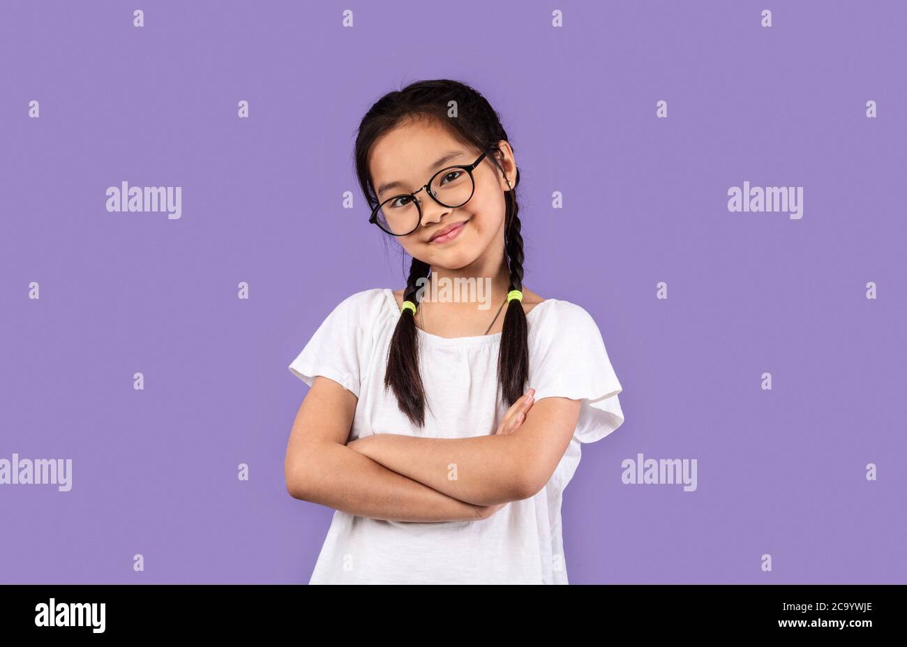 Piccola ragazza cinese che indossa occhiali che posano mani incrociate, sfondo viola Foto Stock