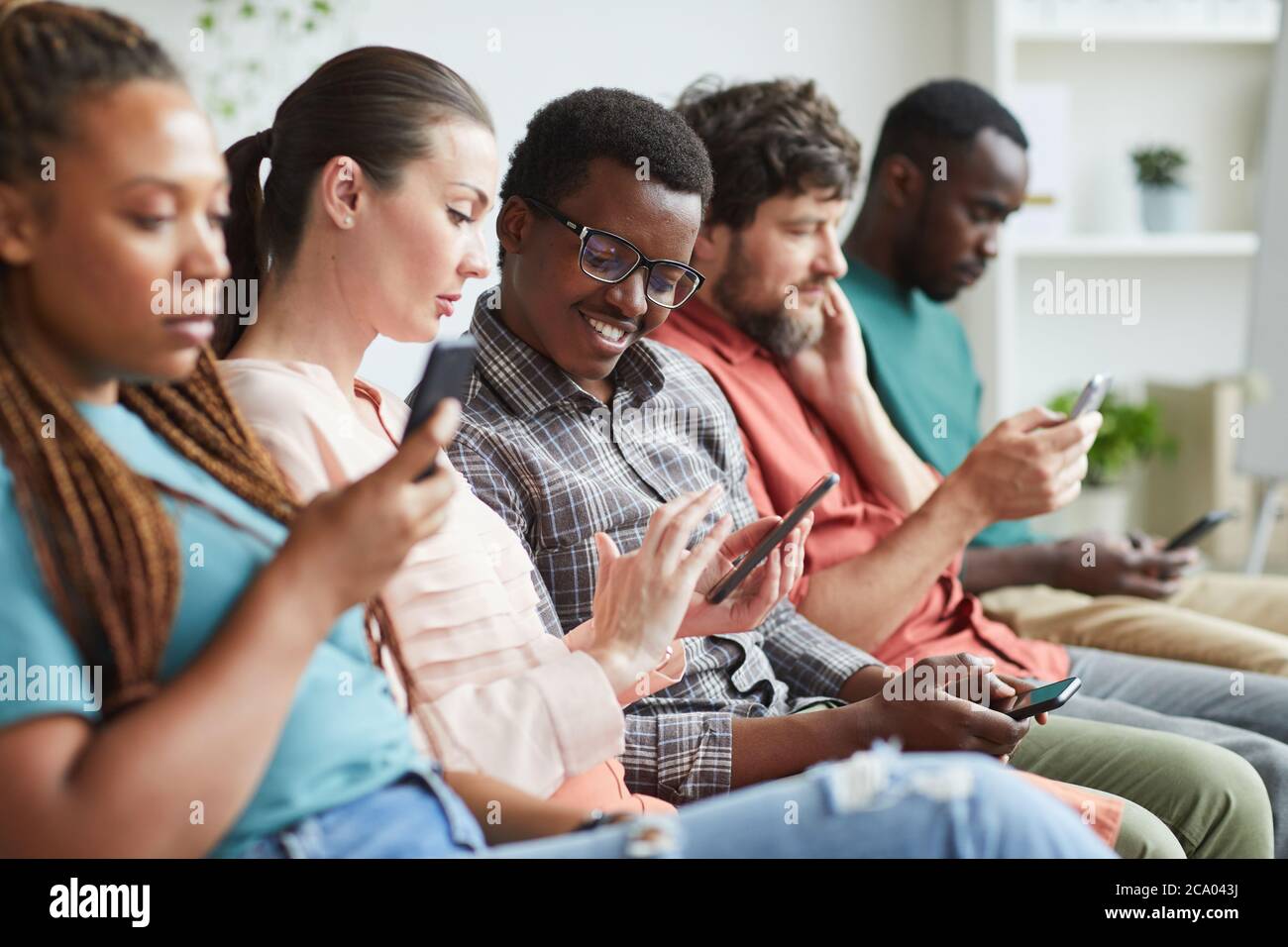 Ritratto di un gruppo multietnico di persone che si siedono in fila e utilizzano gli smartphone in attesa di una conferenza, concentrarsi sul sorriso afroamericano che parla con la collega accanto a lui, copia spazio Foto Stock