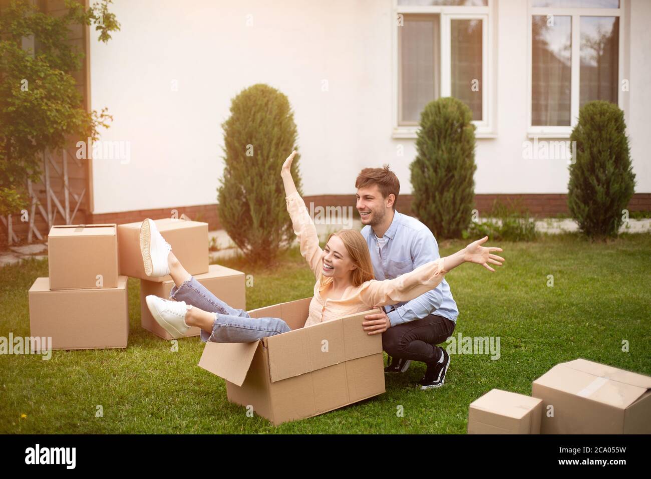 Coppia millennial eccitata che gioca con scatole di cartone vicino alla loro casa di campagna in giorno di movimento Foto Stock