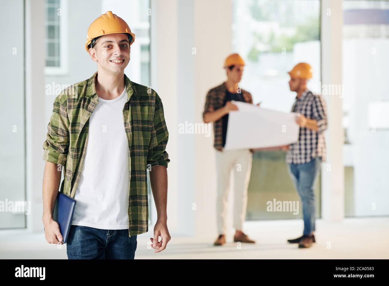Giovane sorridente ingegnere edile medio orientale in camicia plaid che lavora per rifinire gli interni con il suo team Foto Stock