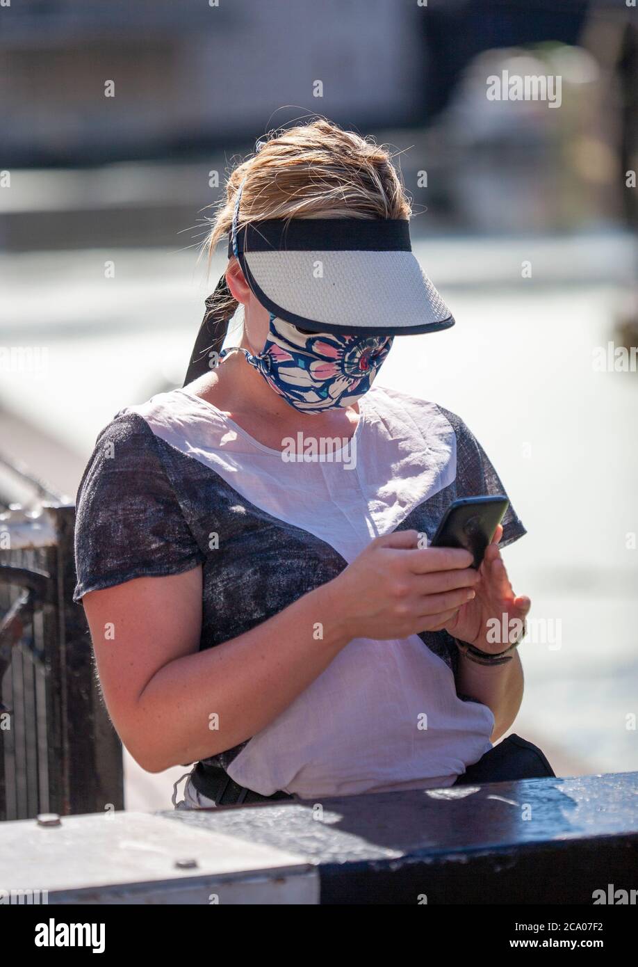 Donna che indossa una visiera parasole e una copertura facciale con motivi utilizzando un telefono cellulare. Londra, Inghilterra Regno Unito Foto Stock