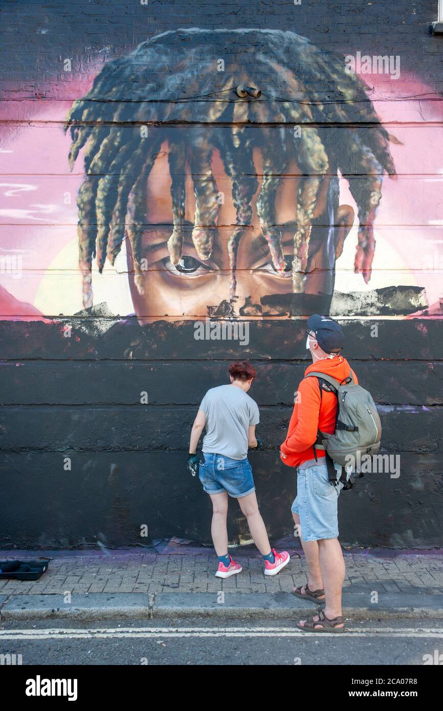 Due persone che dipingano su un murale di graffiti, di un giovane maschio nero con dreadlock, a Camden, Londra, Inghilterra Foto Stock