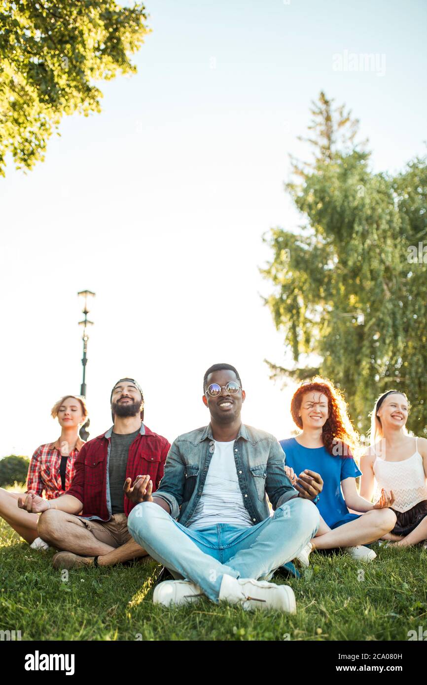Giovane afroamericano che indossa occhiali da sole, che conduce una lezione di yoga in un parco pubblico all'aperto. Amici multirazziali in abiti estivi casual decidere di ha Foto Stock