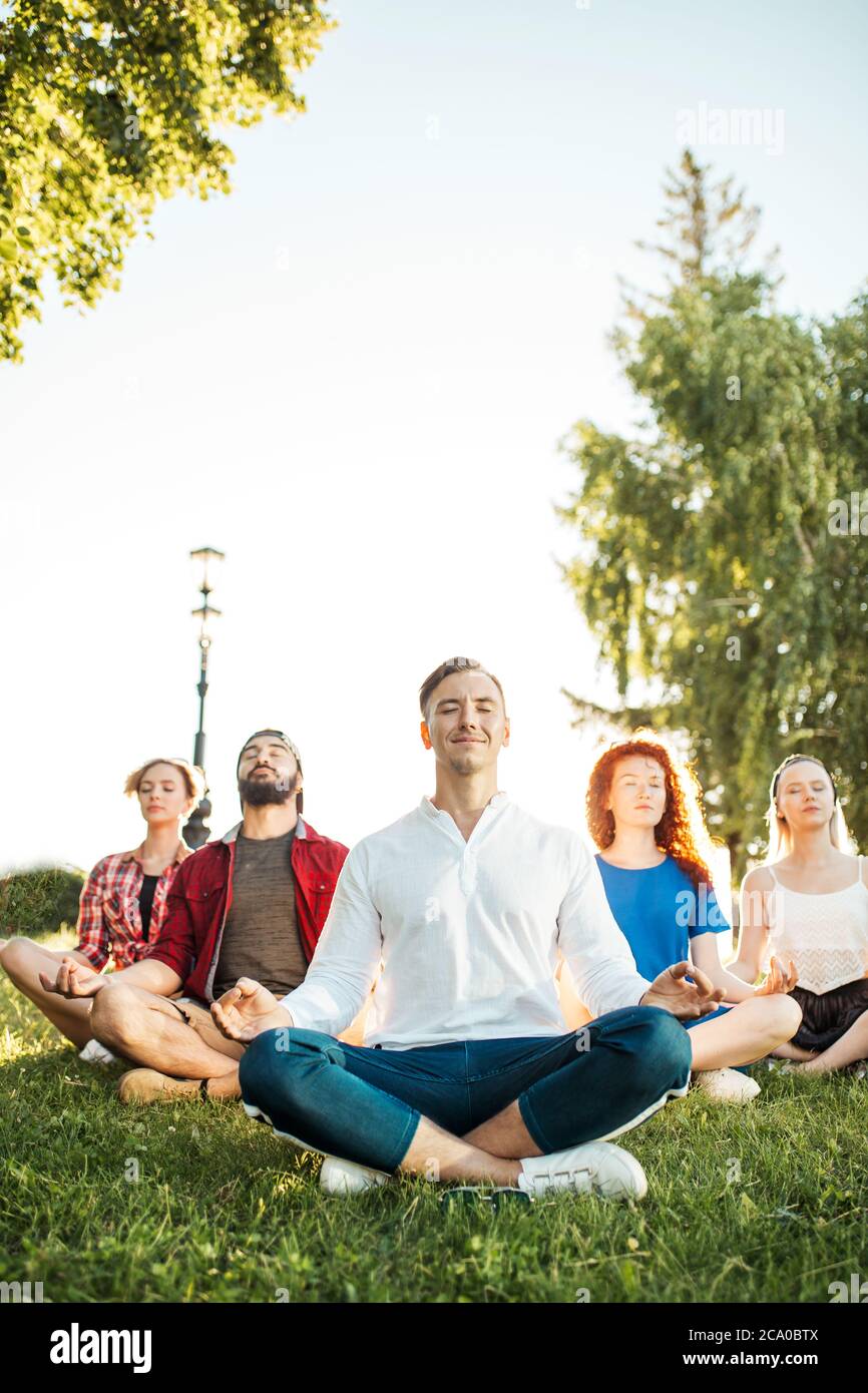 Diverse lezioni di yoga di giovani amici allegri in estate indossare casual seduto in loto posa sul verde nel parco pubblico, vista frontale con messa a fuoco su ma Foto Stock