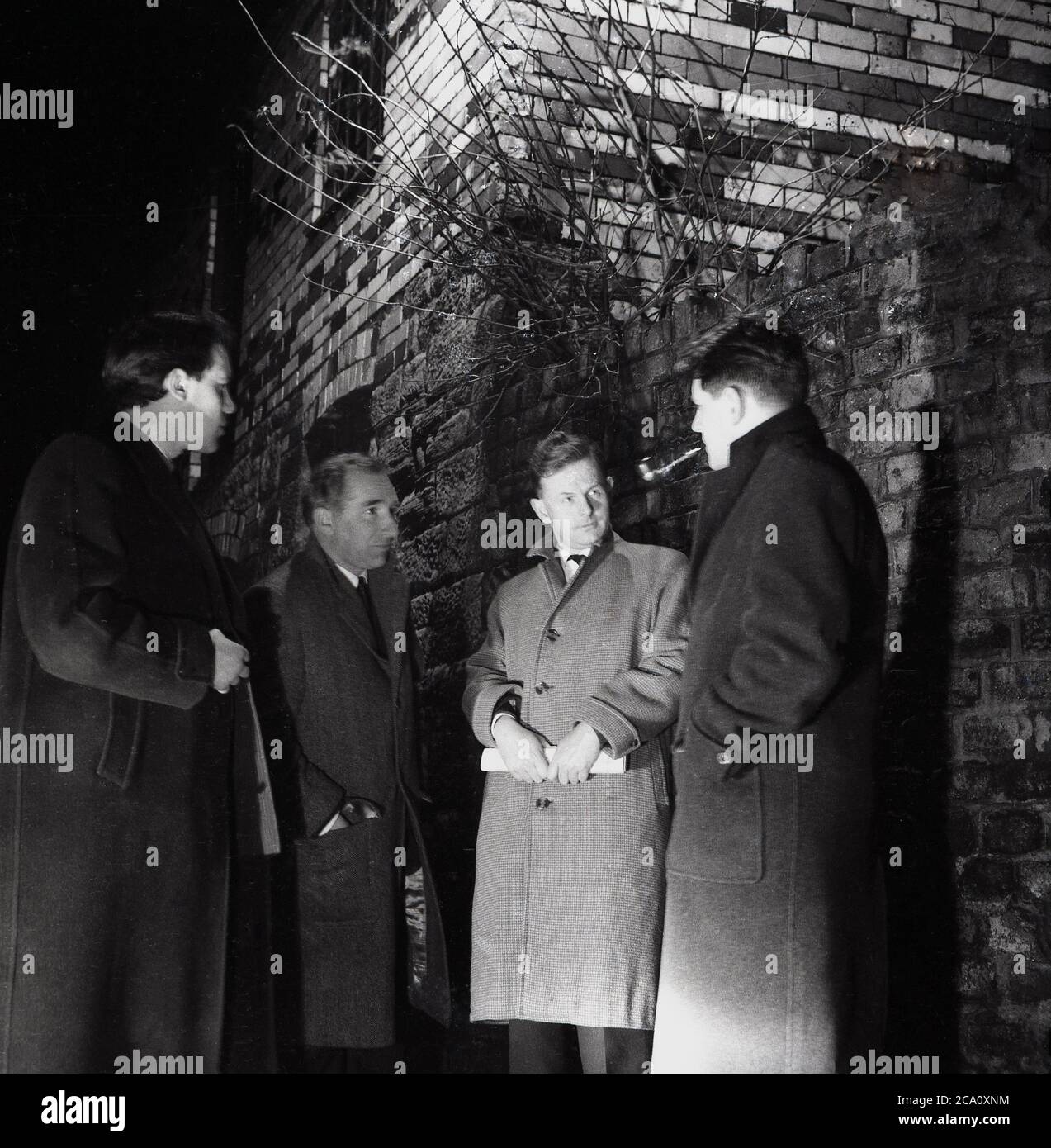 1960, storico, eveningtime e quattro uomini che indossano soprabiti e uno fumare un tubo in piedi in un vicolo che ha un segreto o riunione di clandetine, in modo da non essere sopravuocato o visto da nessuno, Londra, Inghilterra, Regno Unito. Foto Stock