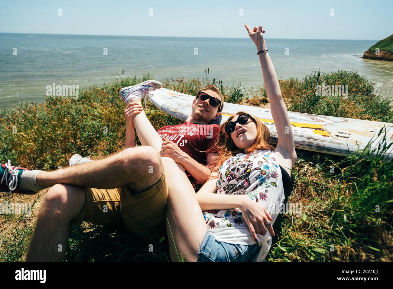 ODESSA, UCRAINA - MAGGIO, 20 2015: Carino giovane coppia hipster di surfisti con surf facendo divertimento e scherzando, sdraiato sull'erba Foto Stock