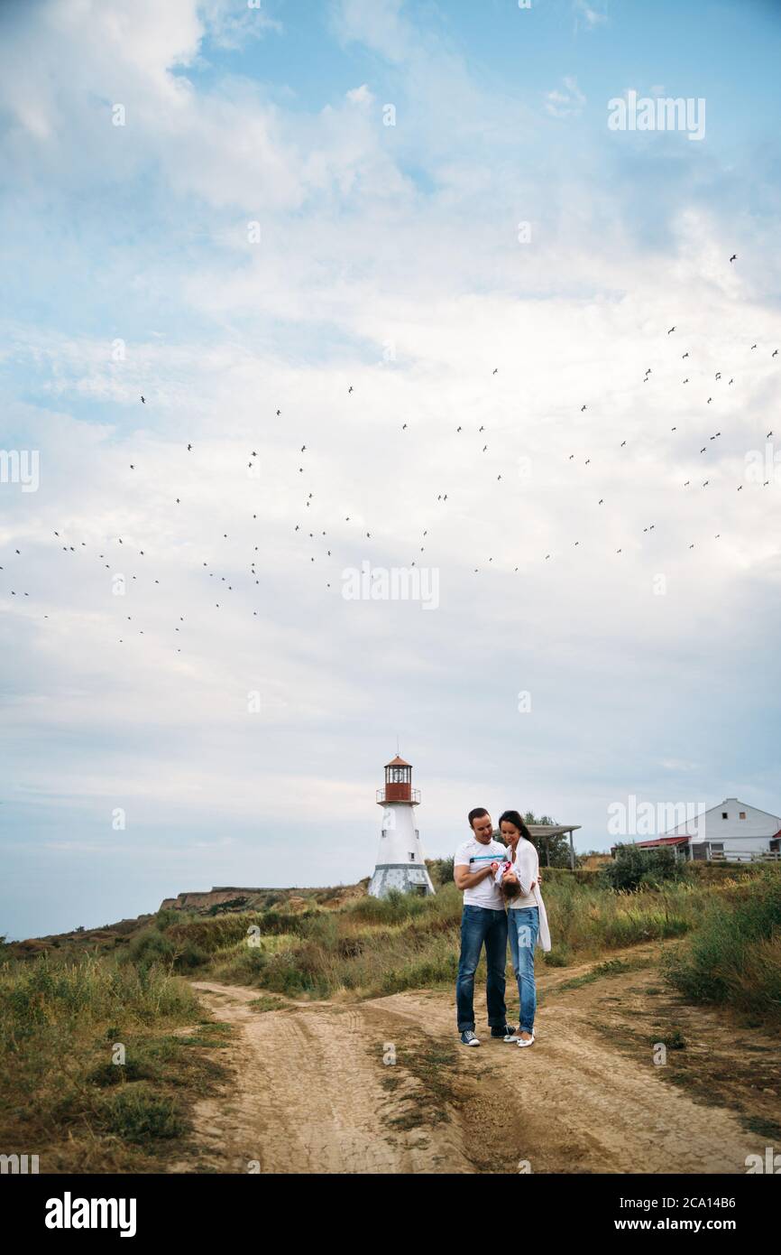 Felice giovane famiglia in magliette bianche e jeans blu con una piccola figlia vicino al faro bianco, sfondo all'aperto con gabbiani nel cielo Foto Stock