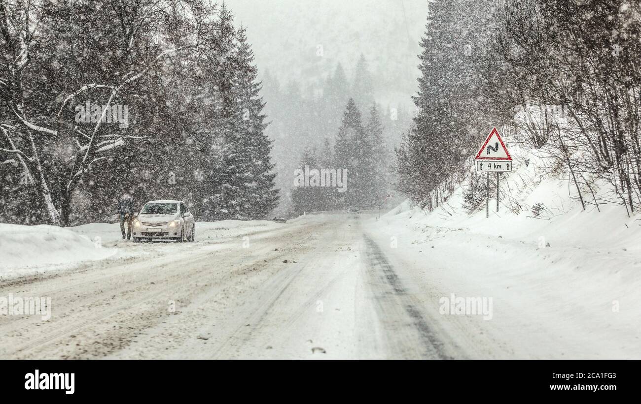 Guida attraverso la tempesta di neve pesante bufera su strada forestale, curve di avvertimento avanti segno a destra, e anonimo uomo con la sua auto ferma sul lato sinistro della strada. Foto Stock