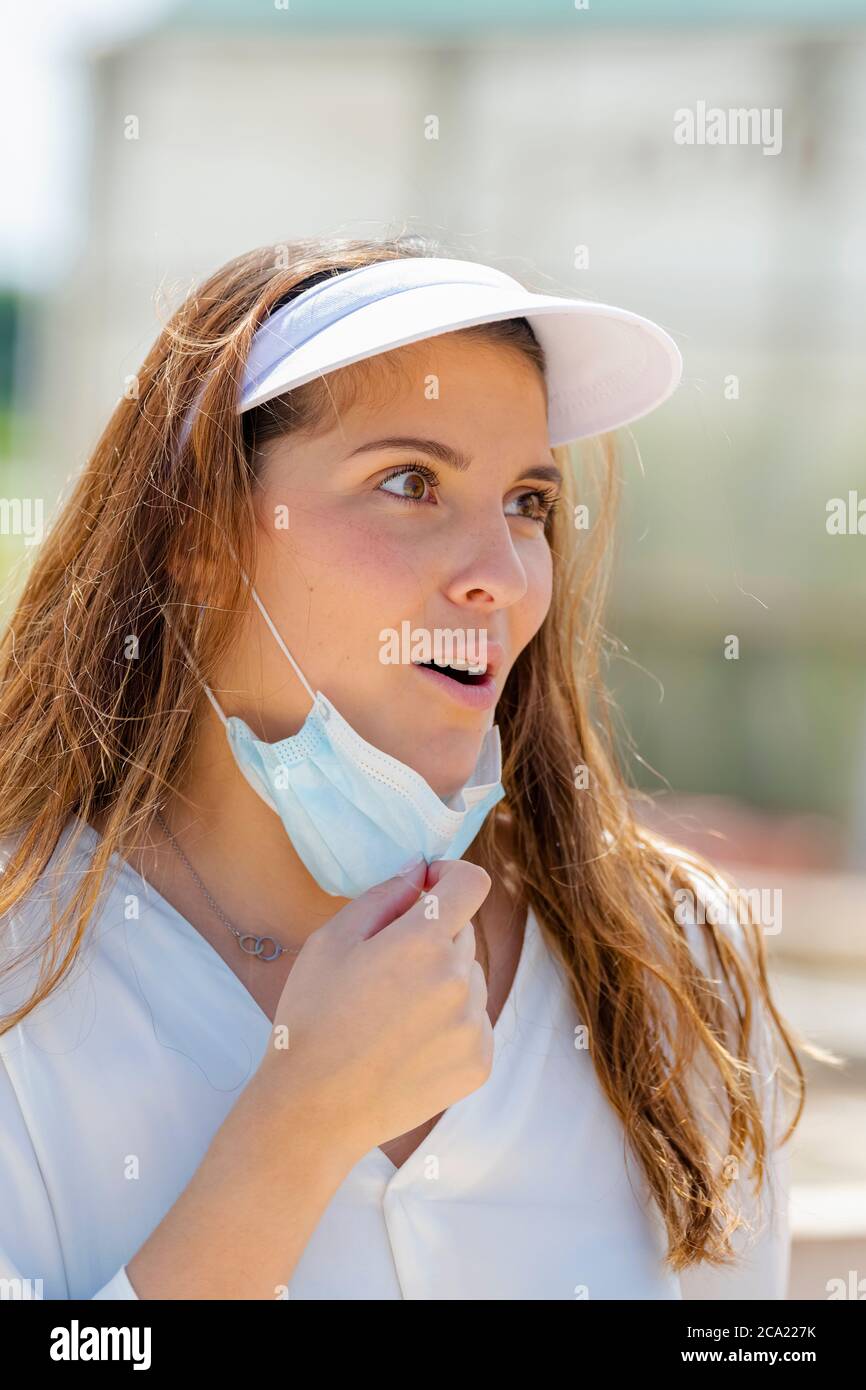 Giovane bella donna che guarda sorpreso mentre tira giù la sua maschera chirurgica su uno sfondo fuori fuoco. Nuovo concetto di stile di vita e sicurezza. Foto Stock