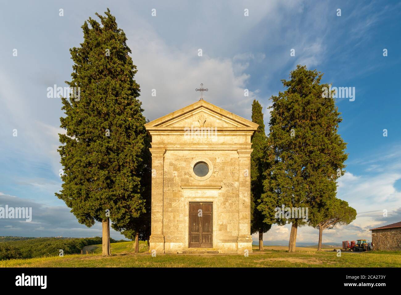 Vista frontale dell'iconica cappella di Vitaleta, in cima alla collina toscano, circondata da cipressi, sotto un cielo blu estivo con nuvole striate Foto Stock