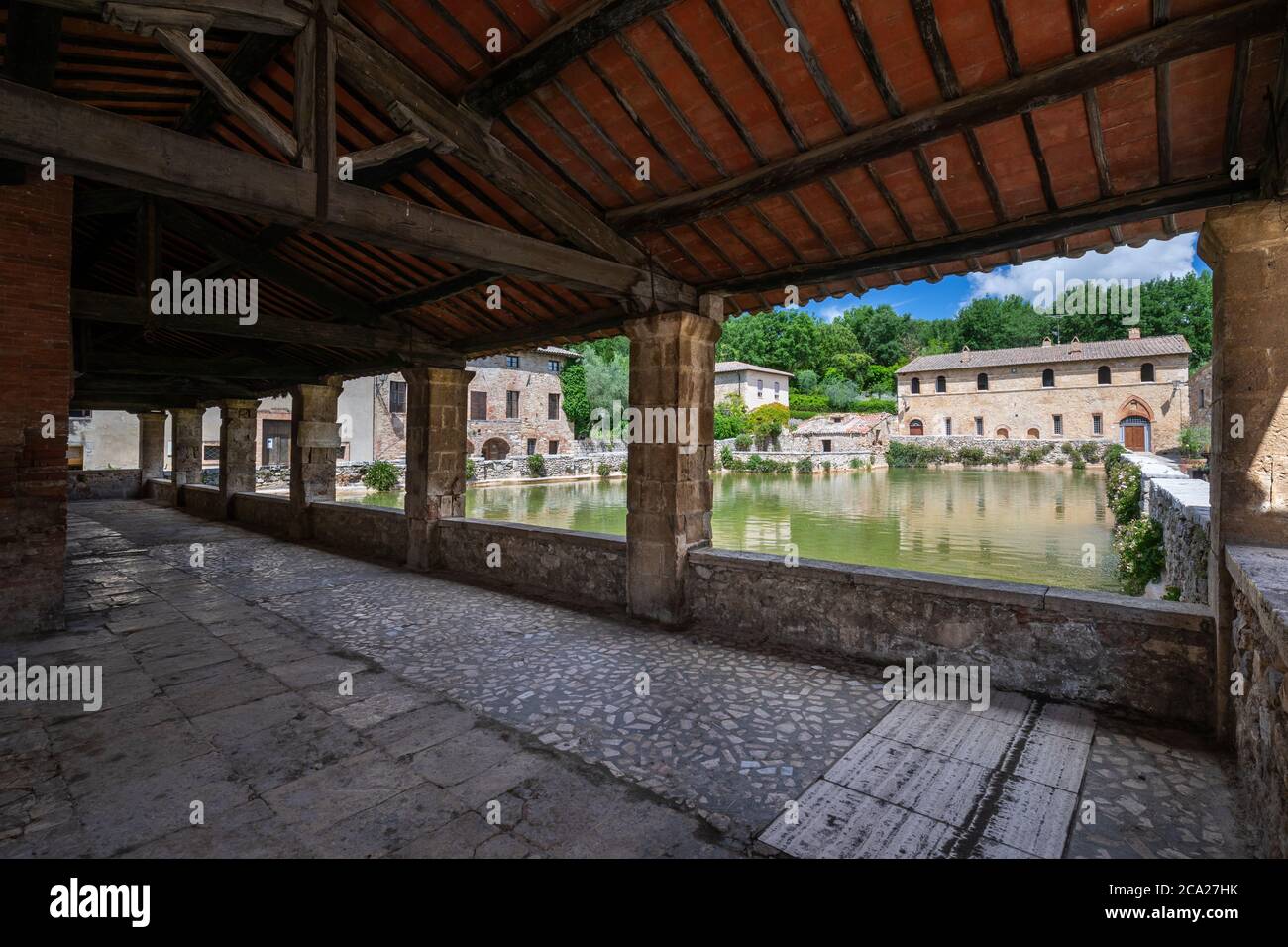 Ampia vista angolare dell'iconica piscina termale medievale di bagno Vignoni, incorniciata da una galleria in pietra e legno, sotto un cielo estivo blu con nuvole soffici Foto Stock