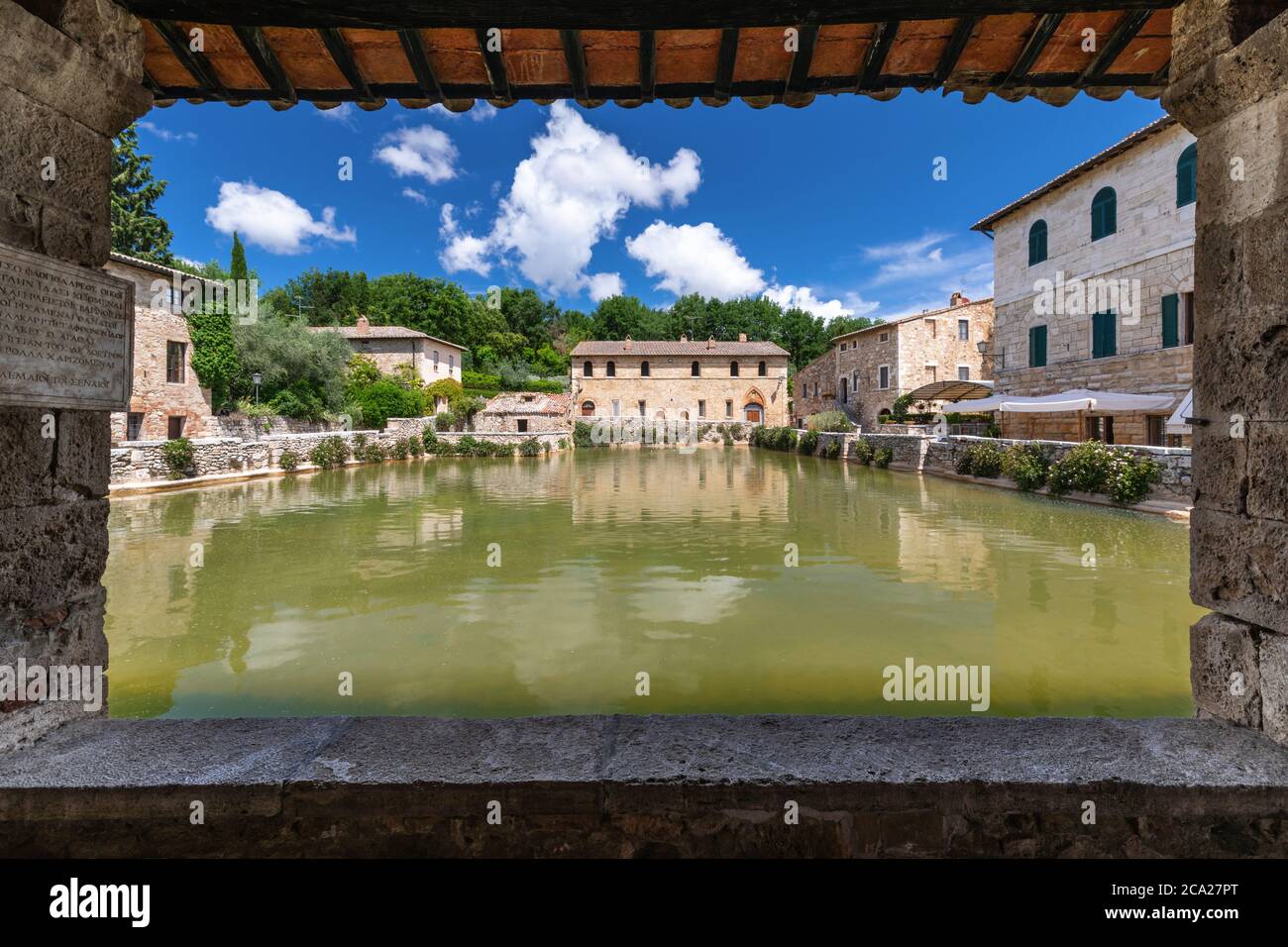 Vista simmetrica dell'iconica piscina termale medievale di bagno Vignoni, incorniciata da una galleria in pietra e legno, sotto un cielo estivo blu con nuvole soffici Foto Stock
