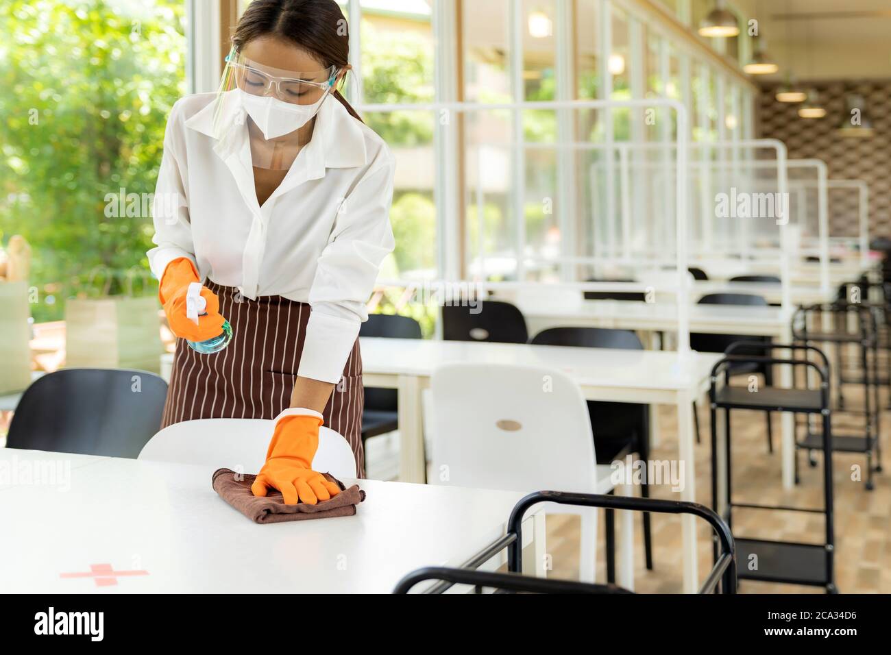 Ritratto attraente cameriera asiatica indossare maschera viso e viso schermo piano di pulizia con alcol e salvietta bagnata prima di accogliere il cliente. Nuovo normale Foto Stock