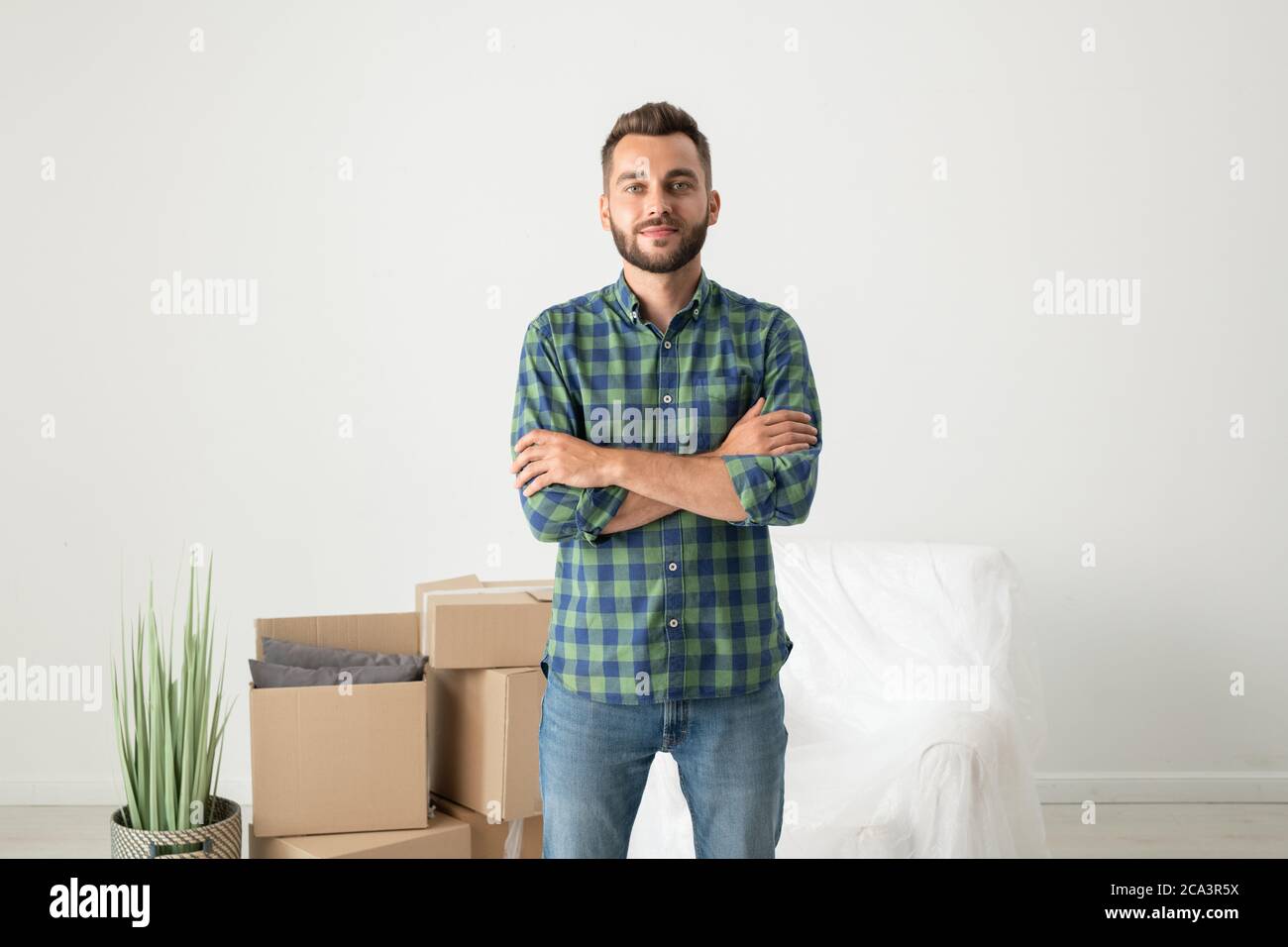 Ritratto di contenuto bel giovane uomo in camicia a scacchi in piedi con braccia incrociate in nuovo piano con scatole mobili Foto Stock
