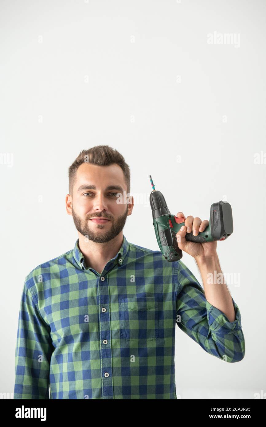 Ritratto del contenuto giovane riparatore con barba in piedi con un cacciavite elettrico contro la parete bianca Foto Stock