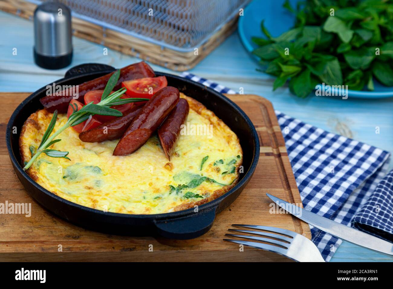 Omelette con salsiccia e pomodori in una padella sul tavolo. Colazione appetitosa. Un rametto di rosmarino, una spezia di legno. Caccia salsicce. Cibo gustoso fritto Foto Stock