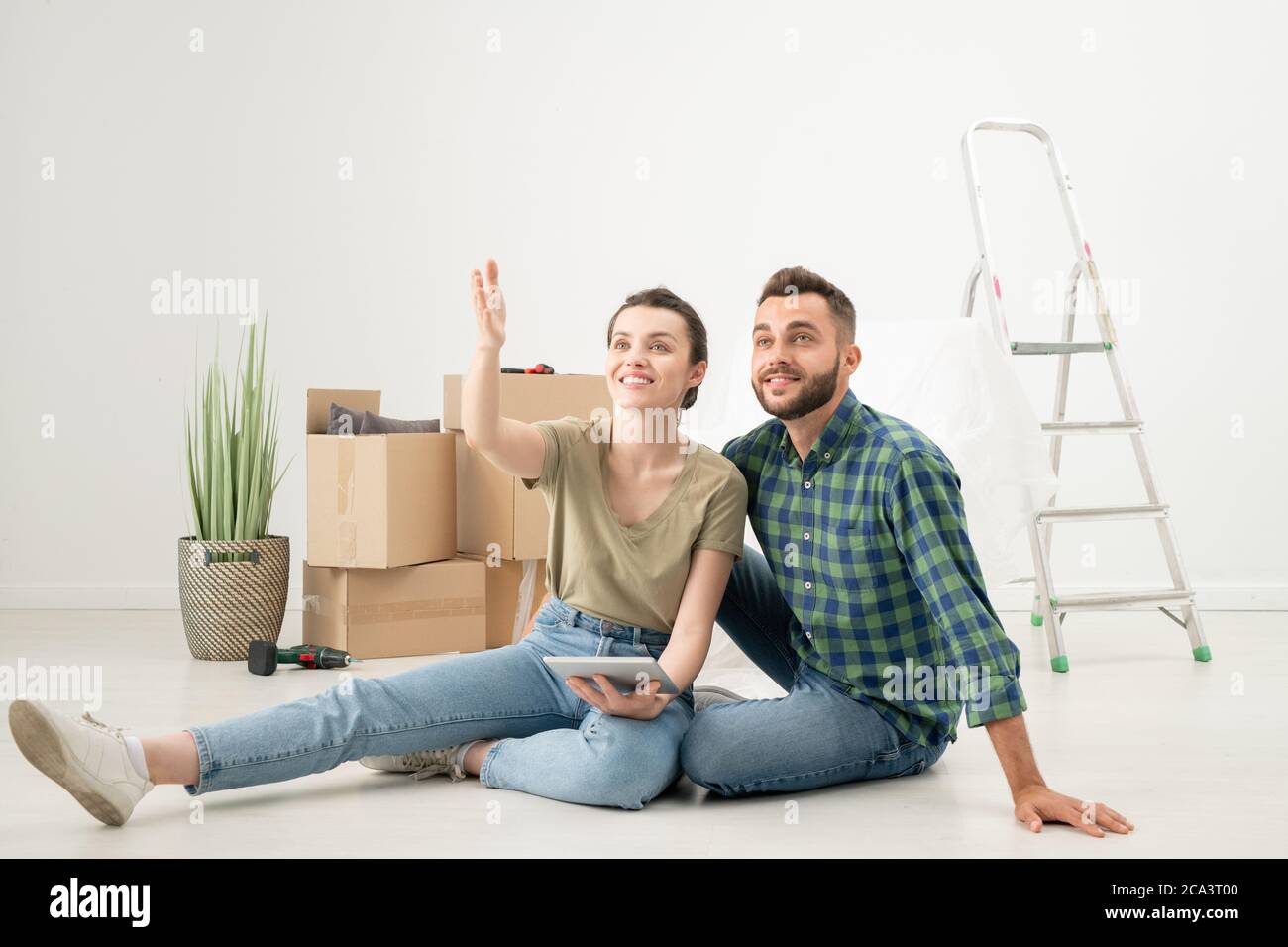 Giovane coppia positiva seduta sul pavimento in un nuovo appartamento con oggetti in movimento e utilizzando l'app di design sul tablet mentre si parla di interni Foto Stock
