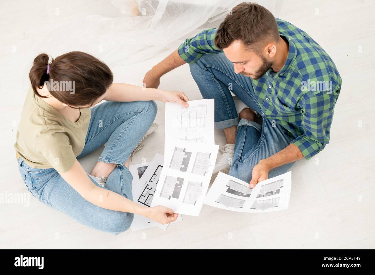 Sopra la vista di giovane coppia concentrata seduta con le gambe incrociate sul pavimento e analizzando i piani della casa mentre si pensa alla ristrutturazione Foto Stock