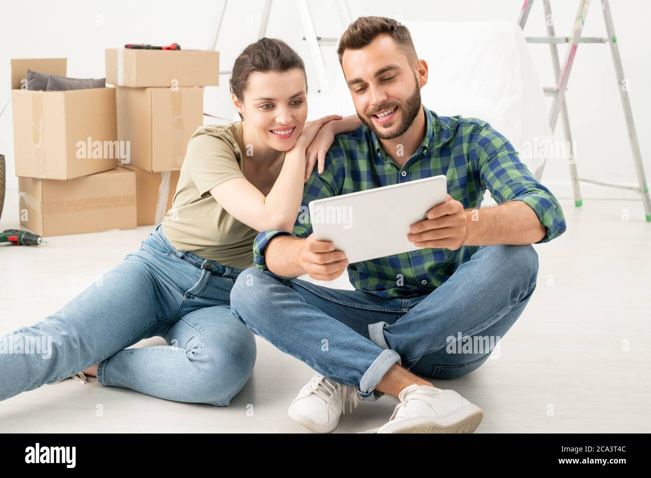 Giovane coppia sorridente seduta sul pavimento in nuovo appartamento e ordinare mobili online utilizzando tablet Foto Stock