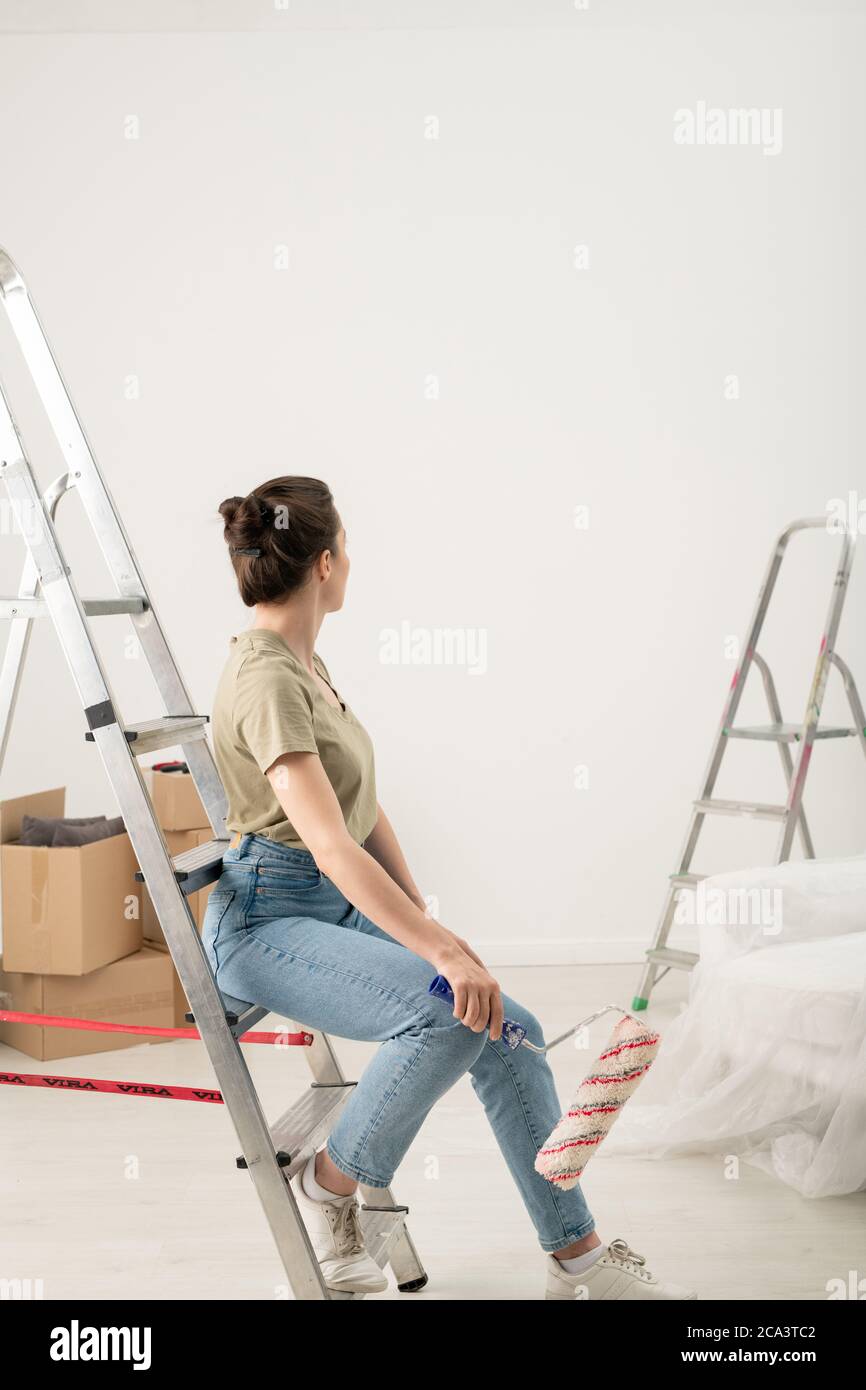 Vista posteriore della donna brunetta in jeans seduta con rullo di vernice sul gradino della scala e guardando la parete dipinta Foto Stock