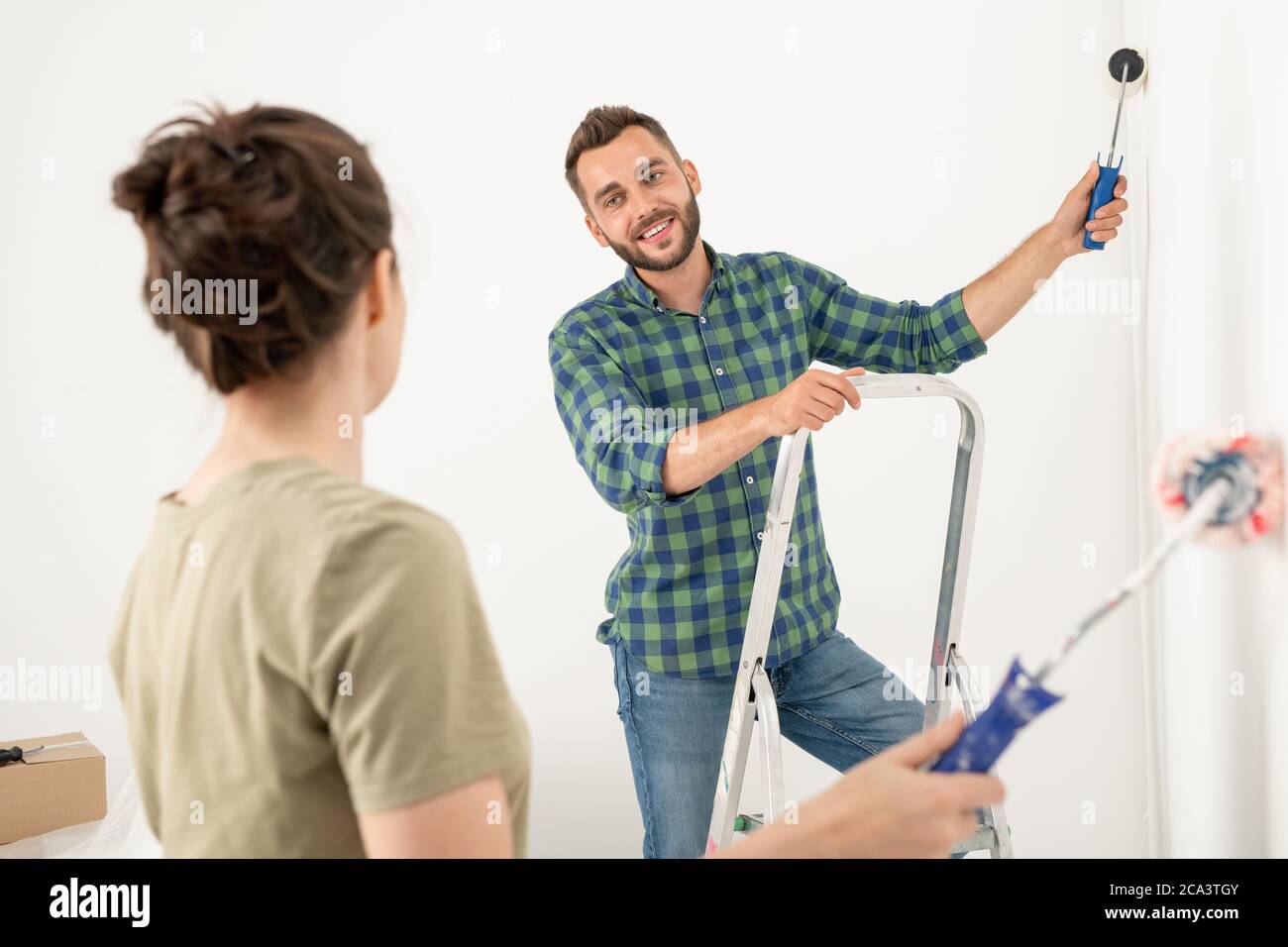 Sorridendo giovane bell'uomo che laccava la parete con un rullo di vernice mentre rimodellava la stanza con la moglie Foto Stock