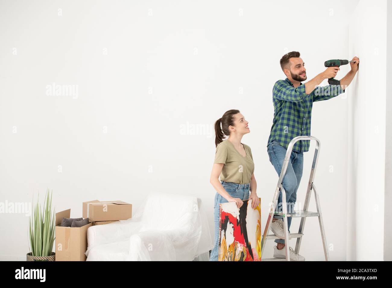 Giovane uomo positivo in piedi sulla scala a gradini e utilizzando un cacciavite elettrico mentre si inserisce la vite per l'immagine Foto Stock