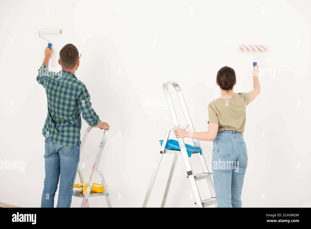 Vista posteriore della giovane coppia dipingere muro bianco utilizzando rullo di vernice durante il rimodellamento della loro vecchia camera Foto Stock